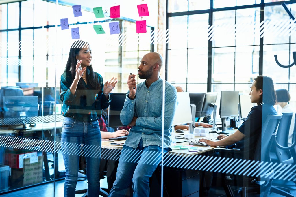 A man perched on the end of an office desk discussing brand strategy with a woman.